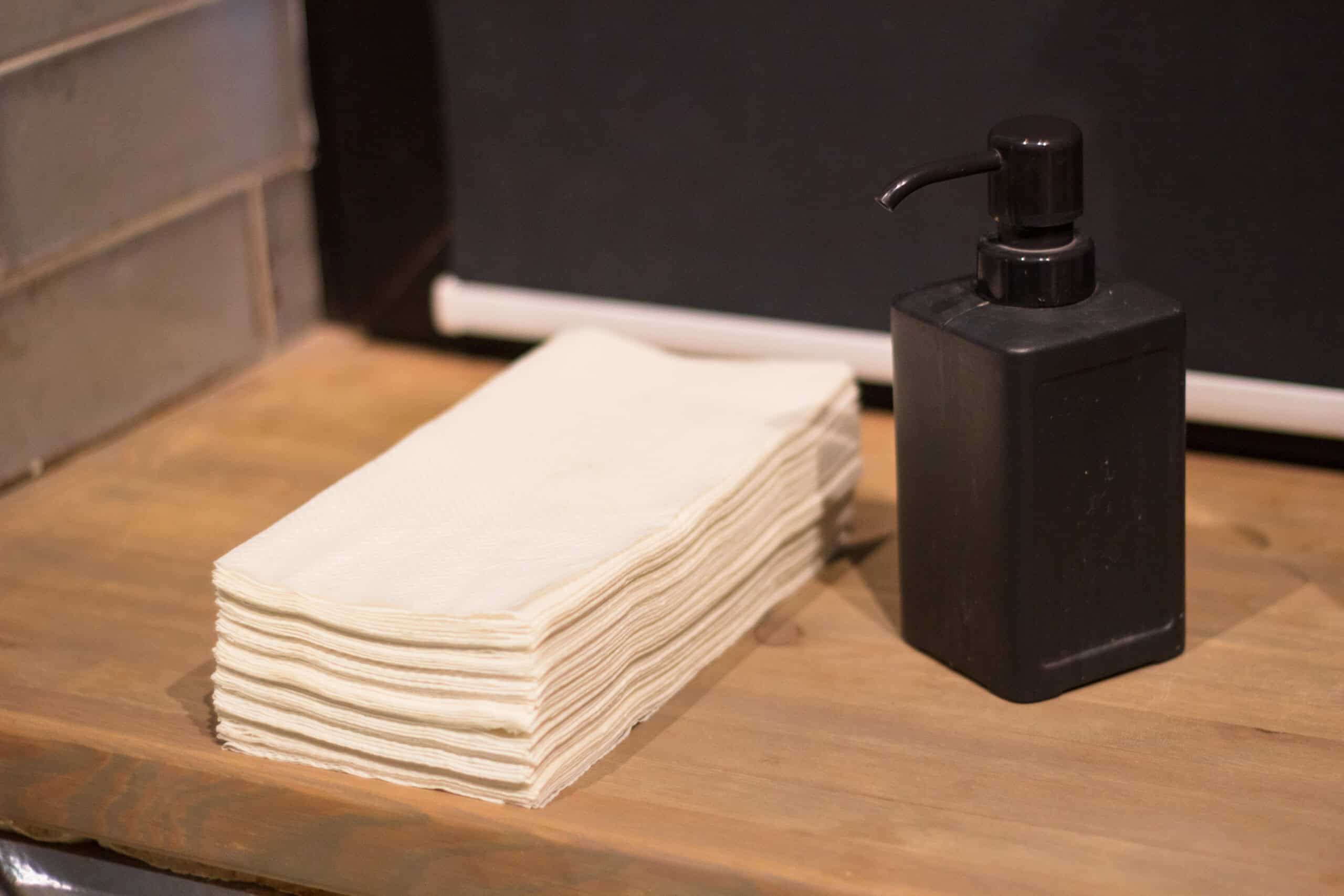 A photo of a stack of paper towels and a black pump bottle on a wooden counter.